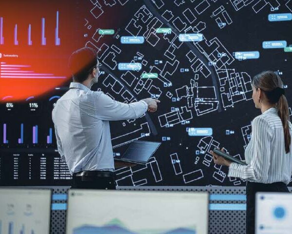 Male And Female Logistics Specialists Discussing New Routs Of Delivery In Front Of Big Digital Screen In Monitoring Office