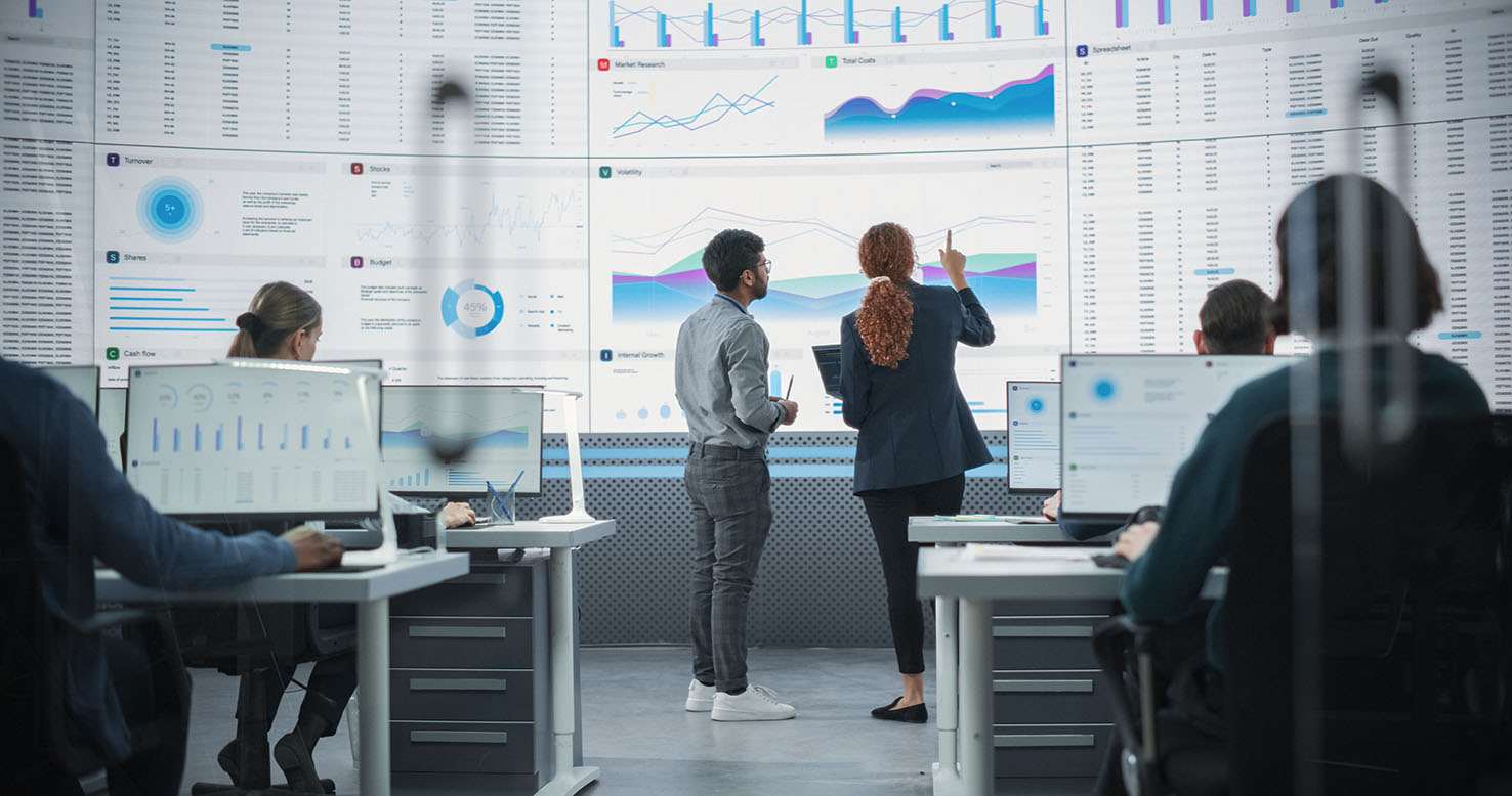 Zoom Out Back View Of Indian Man And Hispanic Woman Discussing Financial Reports On Digital Screen in Monitoring Room. Diverse Colleagues Working On Desktop Computers For Investment Management