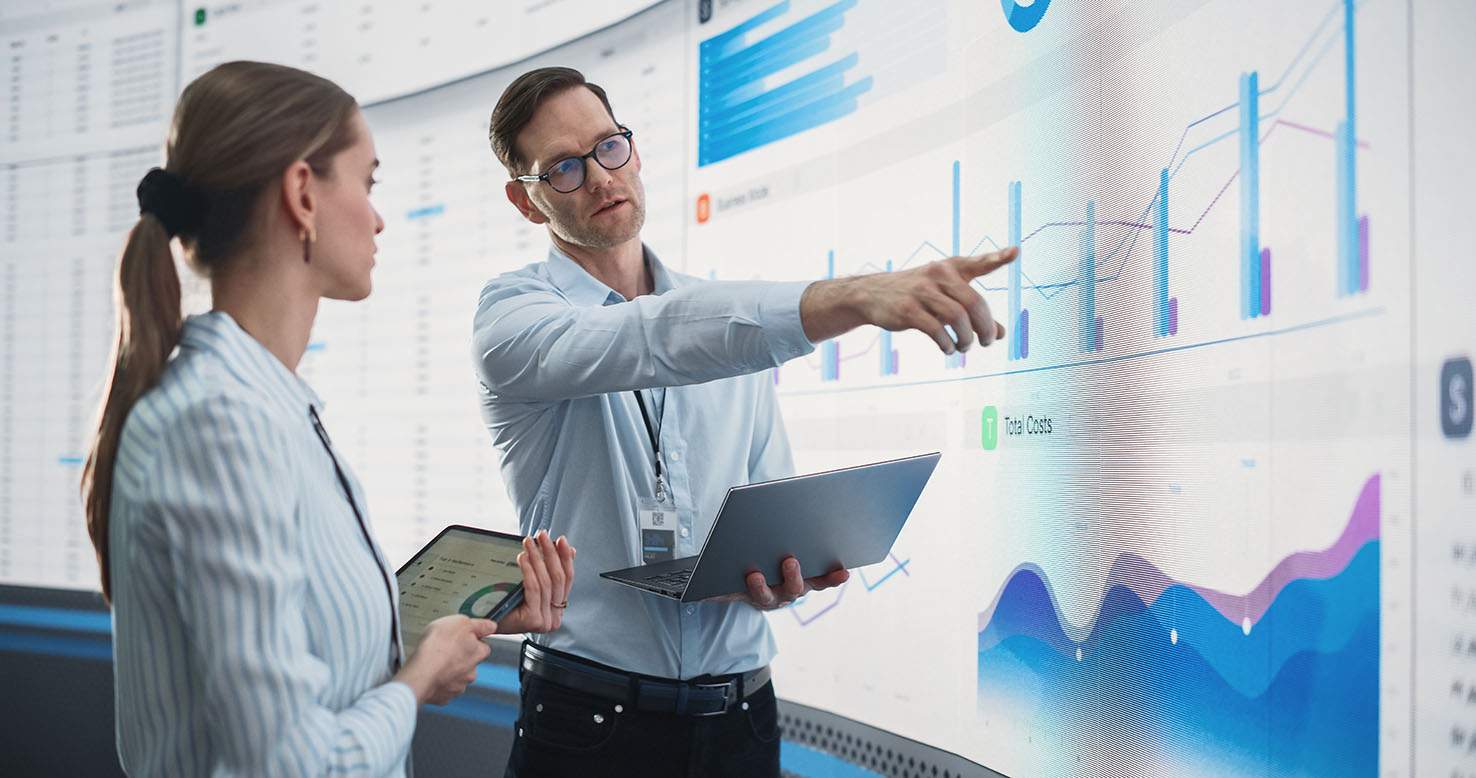 Caucasian Male And Female Data Scientists With Laptop And Tablet Standing Next To Big Digital Screen With Graphs And Charts In Monitoring Office.