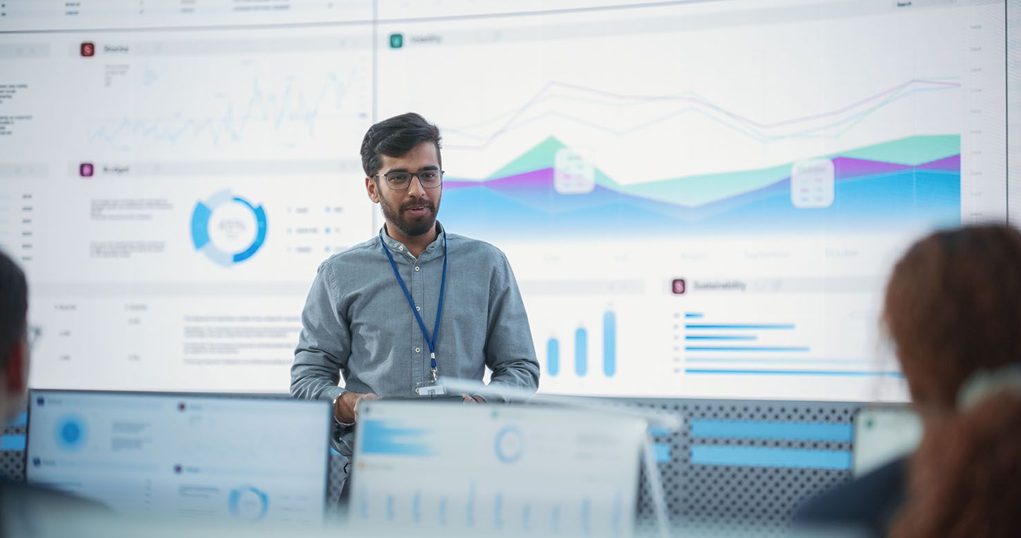 Male Indian Computer Scientist With Tablet Giving Presentation to Diverse Team Of Data Analysts In Front Of Big Digital Screen In Monitoring Room.